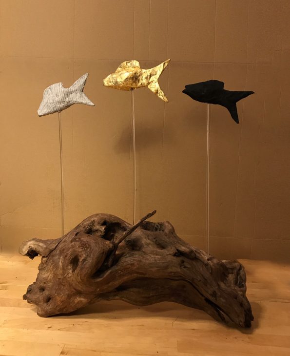 Gebogenes Treibholz, darauf auf Acrylstäben montier drei Fische, links Zeitung, Mitte Blattgold, rechts schwarz